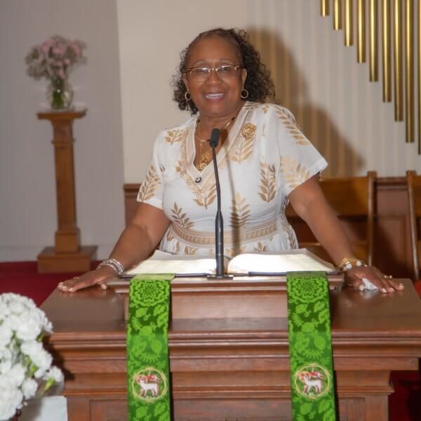 Pastor Blondel Jones-Grant smiles standing behind pulpit during her installation wearing cream dress with gold-leaf print at Great Kills Moravian Church.